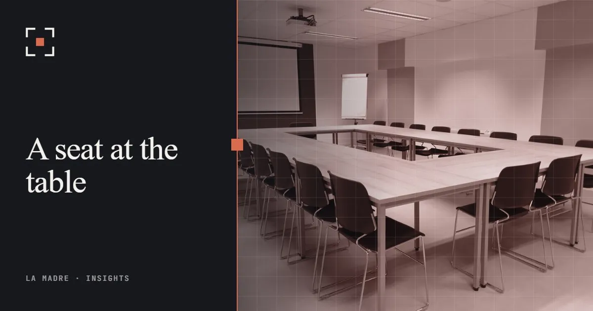 An empty meeting room with tables arranged in a U and chairs around them, in La Madre duotone, beside the words A seat at the table