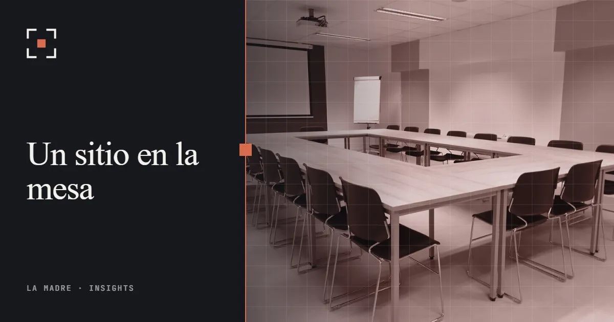 Una sala de reuniones vacía con mesas dispuestas en U y sillas alrededor, en duotono La Madre, junto a las palabras Un sitio en la mesa